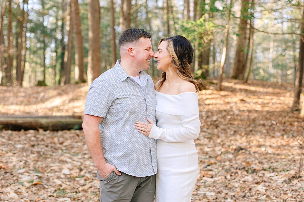 Couple standing close in a forest, touching noses and smiling at each other, surrounded by fallen leaves and tall trees.
