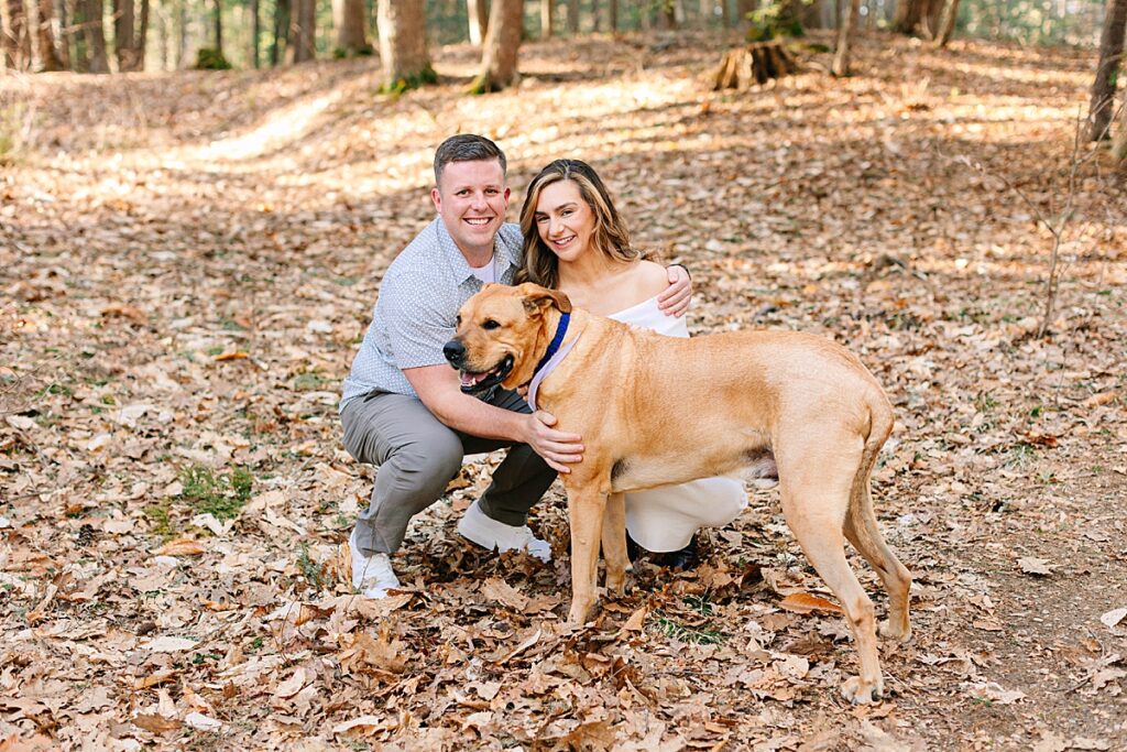 Couple crouching beside their happy tan dog in a forest filled with autumn leaves.