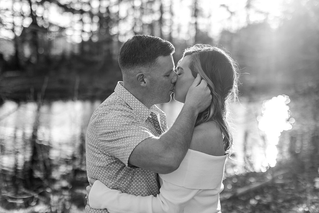 Black and white close-up of a couple kissing near a sunlit pond, framed by soft backlight and trees in the background.