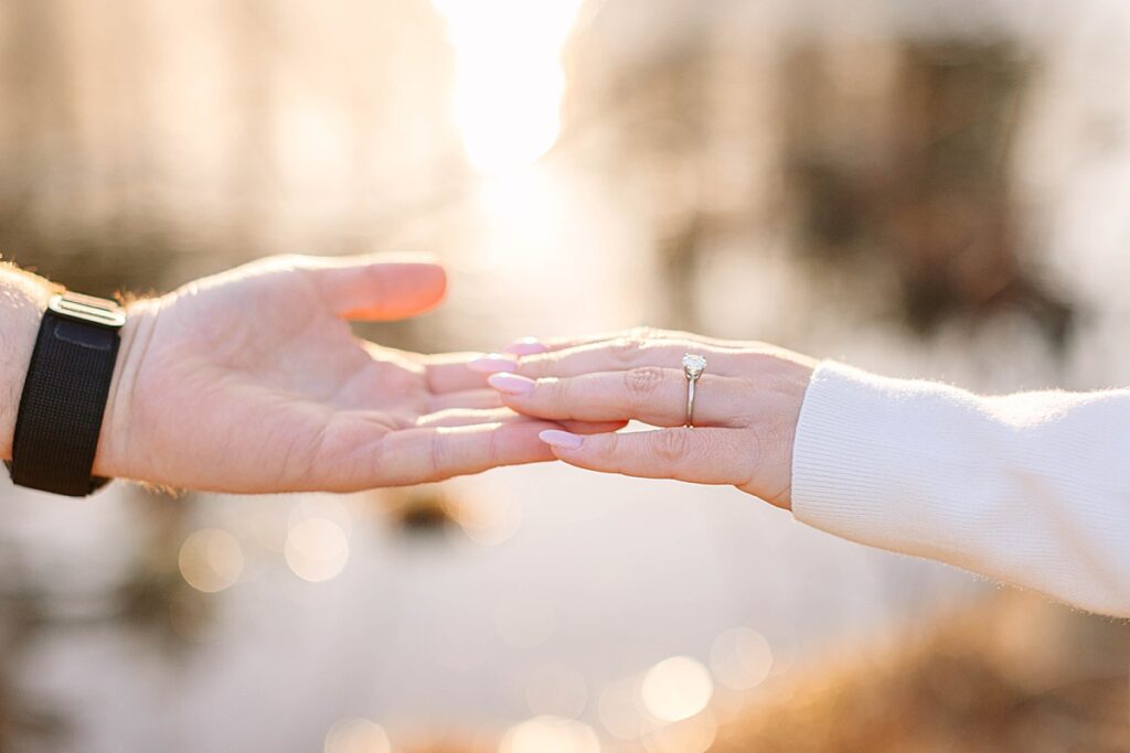 Close-up of hands reaching toward each other, highlighting a round engagement ring with soft sunlight sparkling in the background.