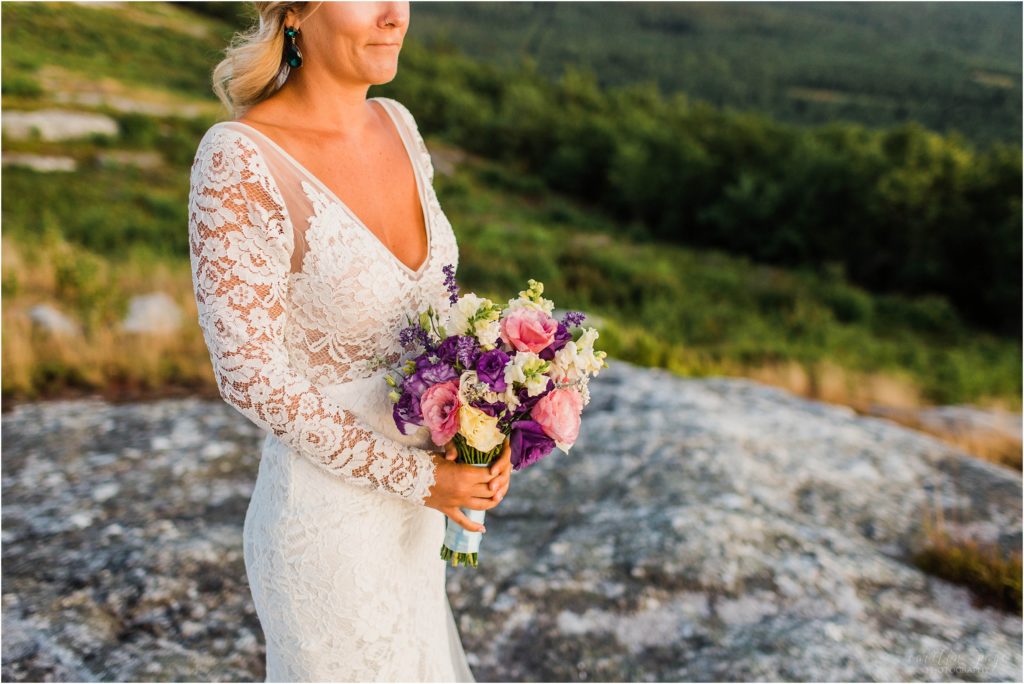 Bride in long sleeve lace dress standing on top of Foss Mountain at sunset with wedding bouquet and long veil