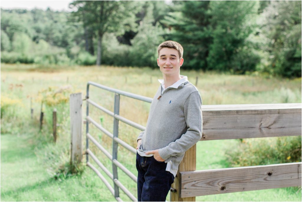 senior boy standing against rail fence in gray sweater