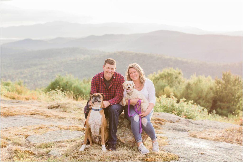 man and woman sitting with two dogs on rock at sunset