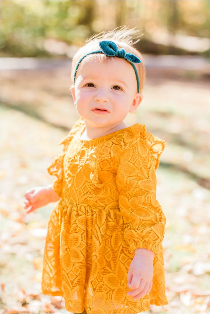 toddler in mustard colored dress standing in yard with leaves