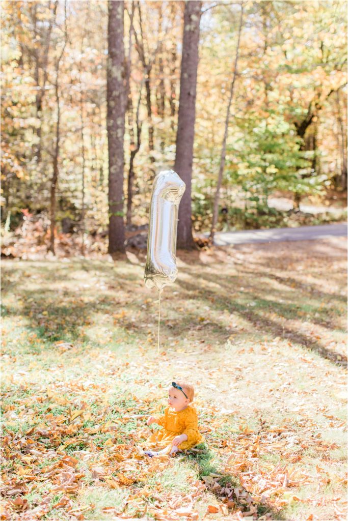 toddler in mustard colored dress sitting in yard with number 1 balloon