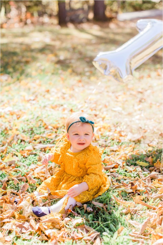 toddler in mustard colored dress sitting in yard with number 1 balloon