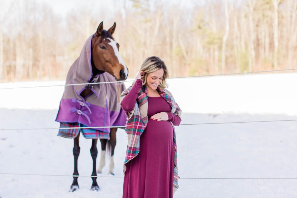 Woman looking at pregnant belly in maternity dress with horse behind her