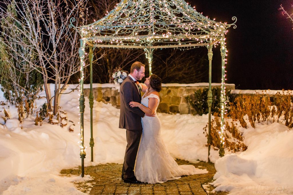 Bride and groom standing under gazebo in the snow with Christmas lights