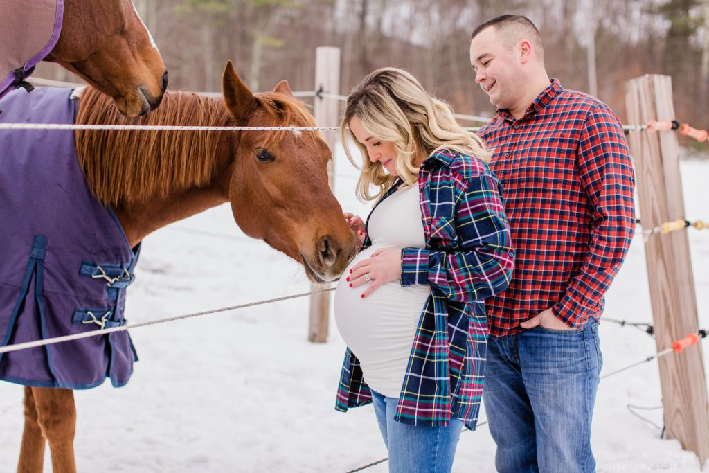 Man and pregnant woman standing in front of a horse that is nuzzling the baby belly