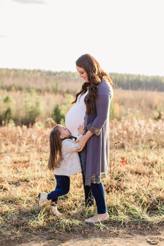 Pregnant woman standing in field while little girl kisses her belly