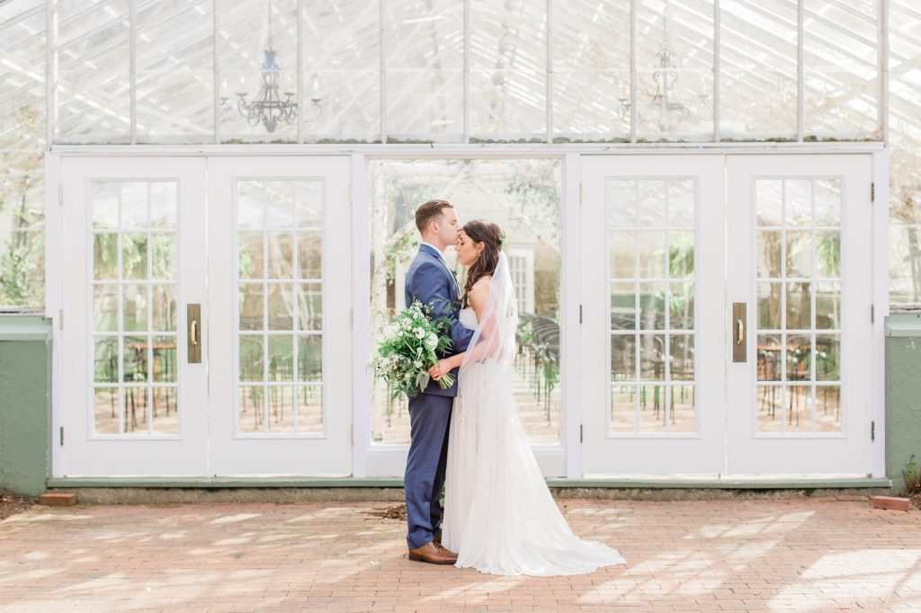 Groom kissing bride on the forehead in front of greenhouse