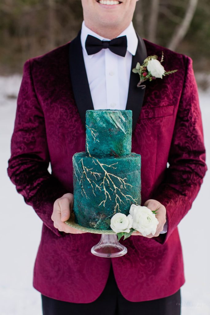 Groom in burgundy tuxedo jacket holding deep turquoise cake with gold details