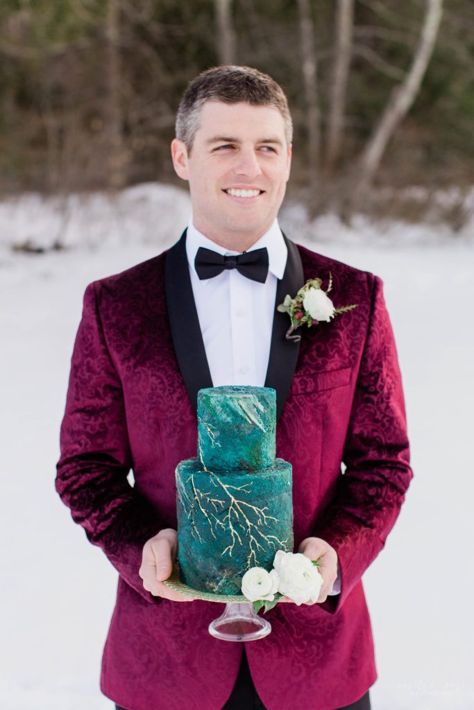 Groom in burgundy tuxedo jacket holding deep turquoise cake with gold details