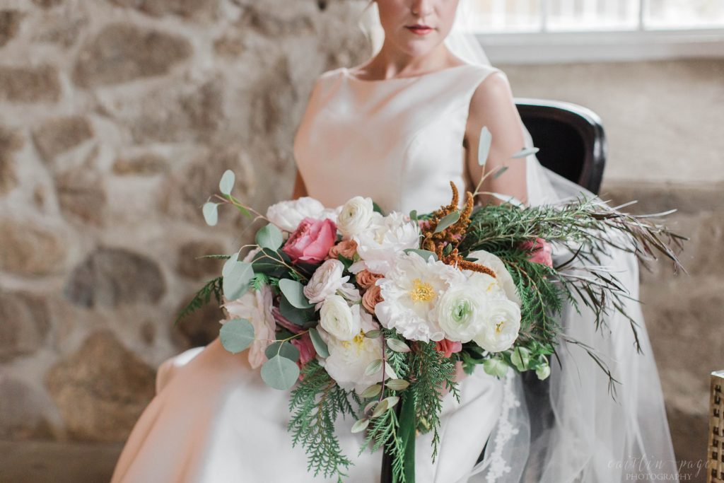 Bride sitting in armchair holding textured wedding bouquet with peonies and greens