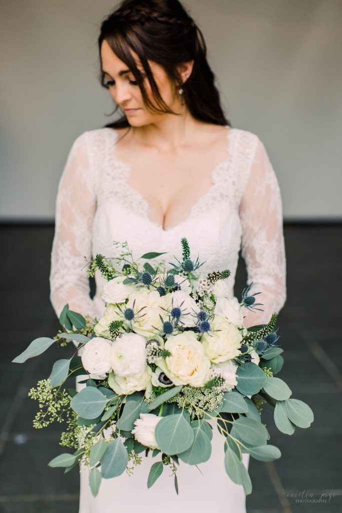 Bride in long sleeved wedding dress holding textured wedding bouquet with blue thistle and greens