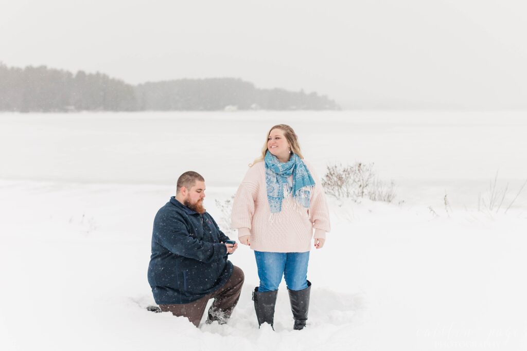 Man on one knee in the snow behind girlfriend