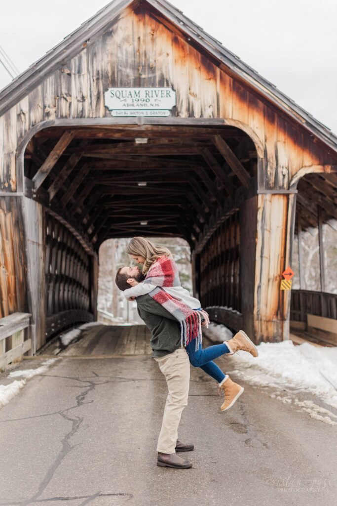 Man holding woman in the air in front of covered bridge