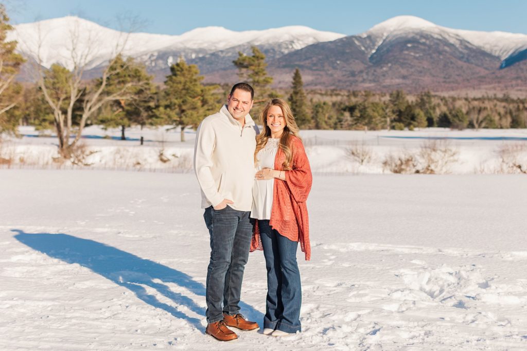 Pregnant woman standing with husband in the snow in front of the mountains