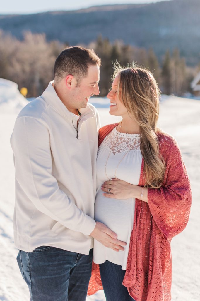 Pregnant woman standing with husband in the snow in front of the mountains