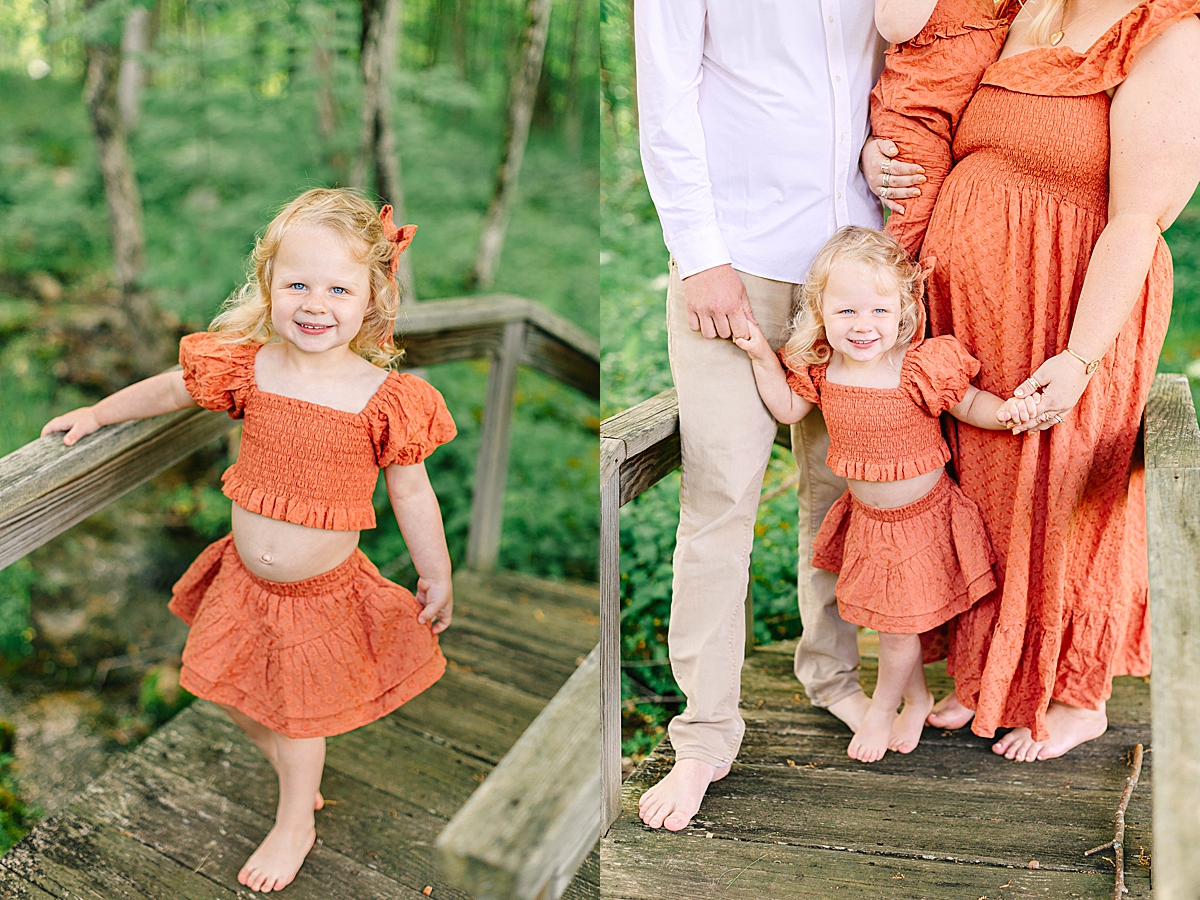 Young girl in an orange two-piece outfit smiling on a wooden bridge in a lush forest. Little girl holding hands with her parents on a bridge, with mom wearing a matching orange dress.