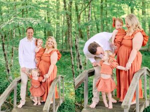 Family of four standing barefoot on a bridge surrounded by green forest, dressed in coordinated outfits. Father leaning down to kiss daughter while mother holds baby on a forest bridge.
