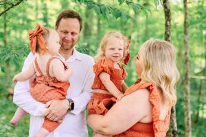 Father holding baby girl while mother carries toddler in matching dresses, under green trees.