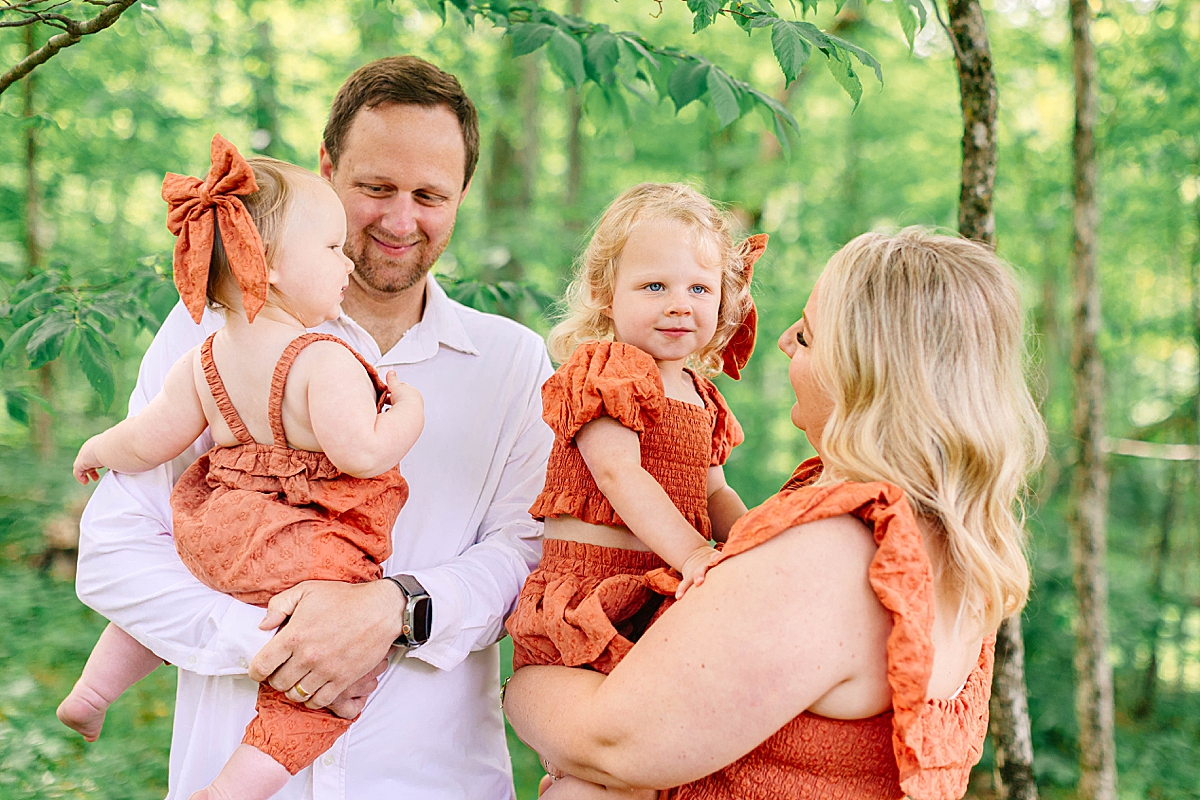 Father holding baby girl while mother carries toddler in matching dresses, under green trees.