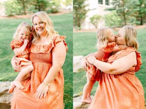Mom sitting on a rock holding her laughing daughter, both wearing coordinating orange dresses. Young girl giving her mom a kiss on the lips while sitting on her lap in a grassy yard.