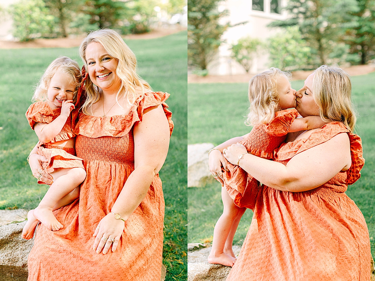 Mom sitting on a rock holding her laughing daughter, both wearing coordinating orange dresses. Young girl giving her mom a kiss on the lips while sitting on her lap in a grassy yard.