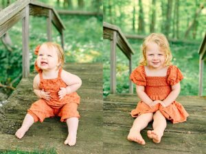 Baby girl in an orange romper sitting on a wooden bridge, smiling with teeth showing. Toddler girl sitting on a forest bridge, smiling with hands in her lap and bare feet outstretched.