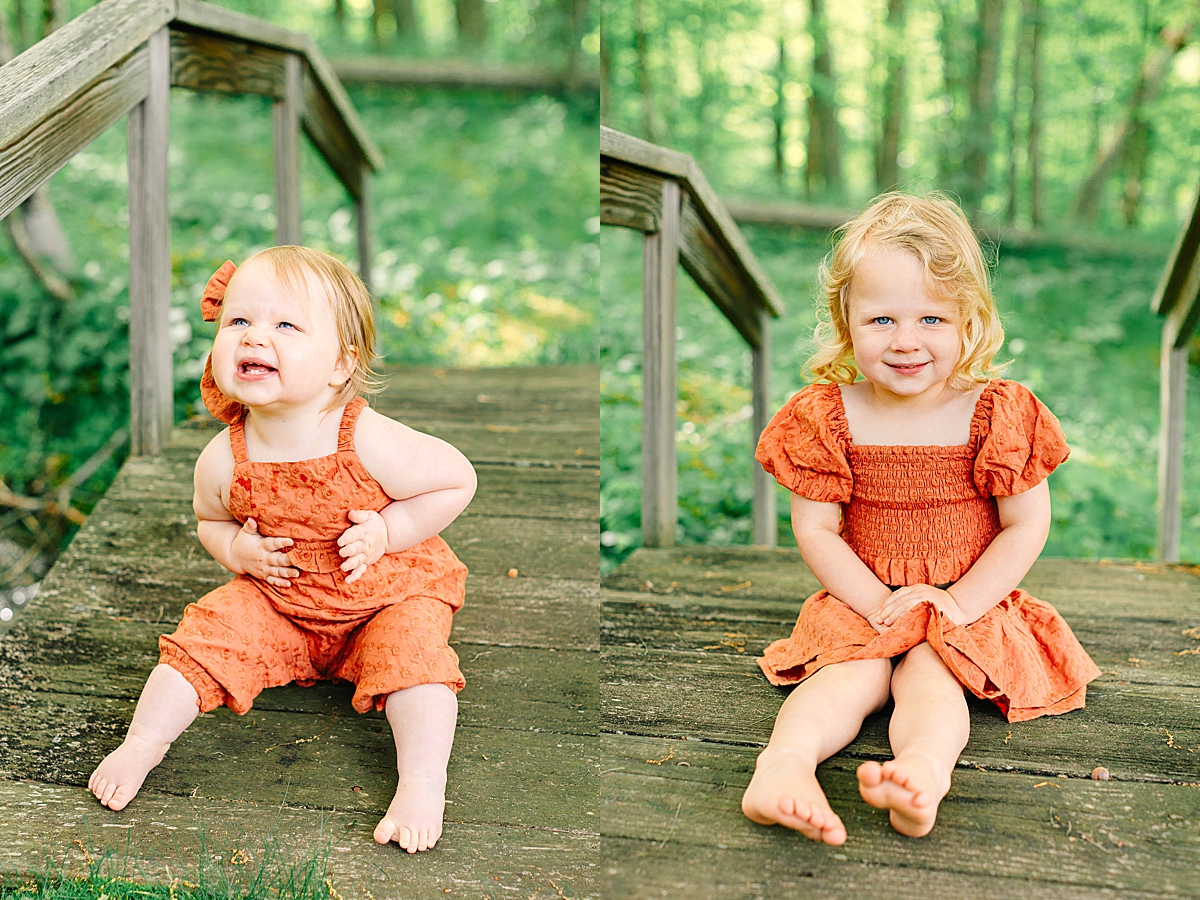 Baby girl in an orange romper sitting on a wooden bridge, smiling with teeth showing. Toddler girl sitting on a forest bridge, smiling with hands in her lap and bare feet outstretched.