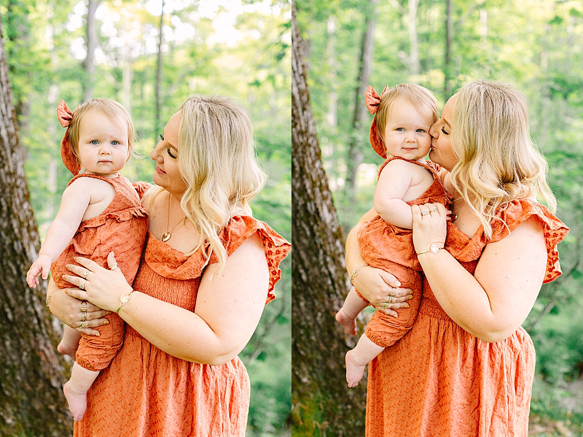 Mom holding baby daughter in the woods, both wearing matching orange tones. Mom kissing baby girl’s cheek while holding her close against a tree-lined background.