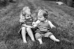 Black and white photo of two young sisters sitting in the grass, gently holding hands and looking at each other.