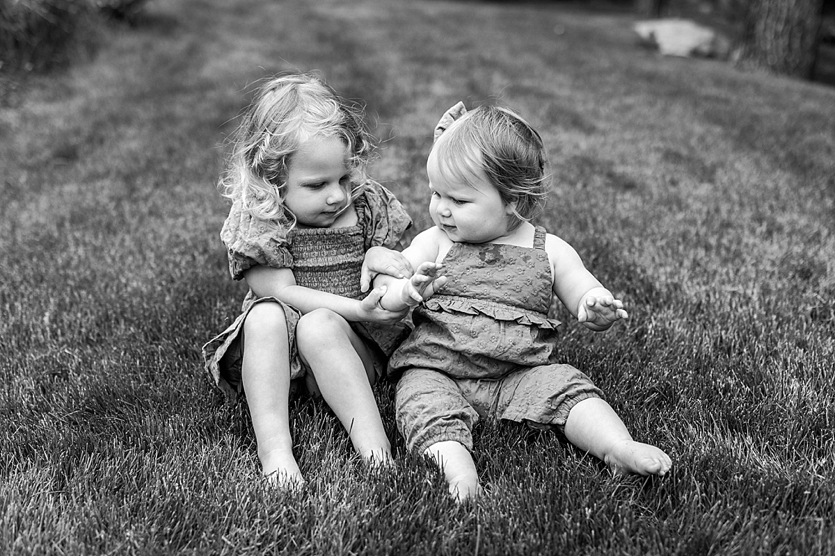 Black and white photo of two young sisters sitting in the grass, gently holding hands and looking at each other.