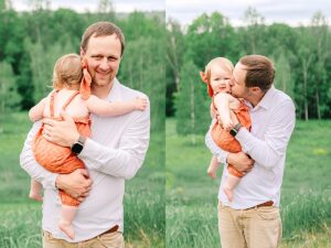 Father holding baby girl on his chest while smiling at the camera, with green trees in the background. Dad kissing baby girl on the cheek while holding her in his arms, standing in a grassy field.