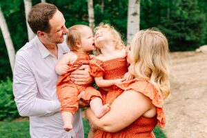 Mom and dad smiling while holding their daughters close, baby kissing toddler on the cheek.