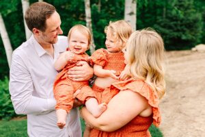 Baby girl smiling in her dad’s arms, with toddler and mom looking on happily.