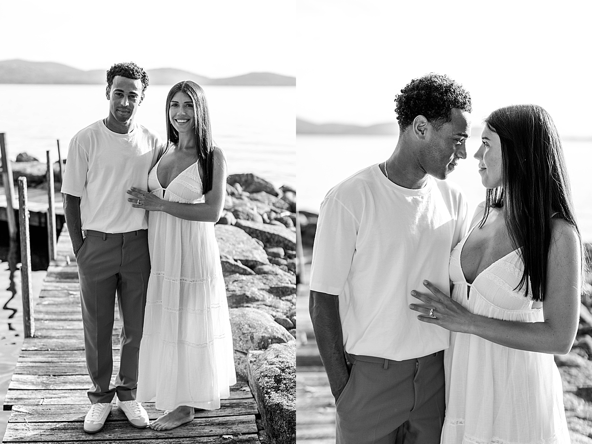 Black and white portrait of a couple standing barefoot on a dock, with a calm lake and mountains in the background.