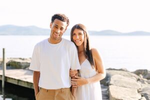 Smiling couple standing arm in arm on a lakeside dock, with soft morning light and mountains in the distance.