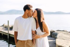 Couple sharing a kiss while standing on a wooden dock, with lake water and hills in the background.