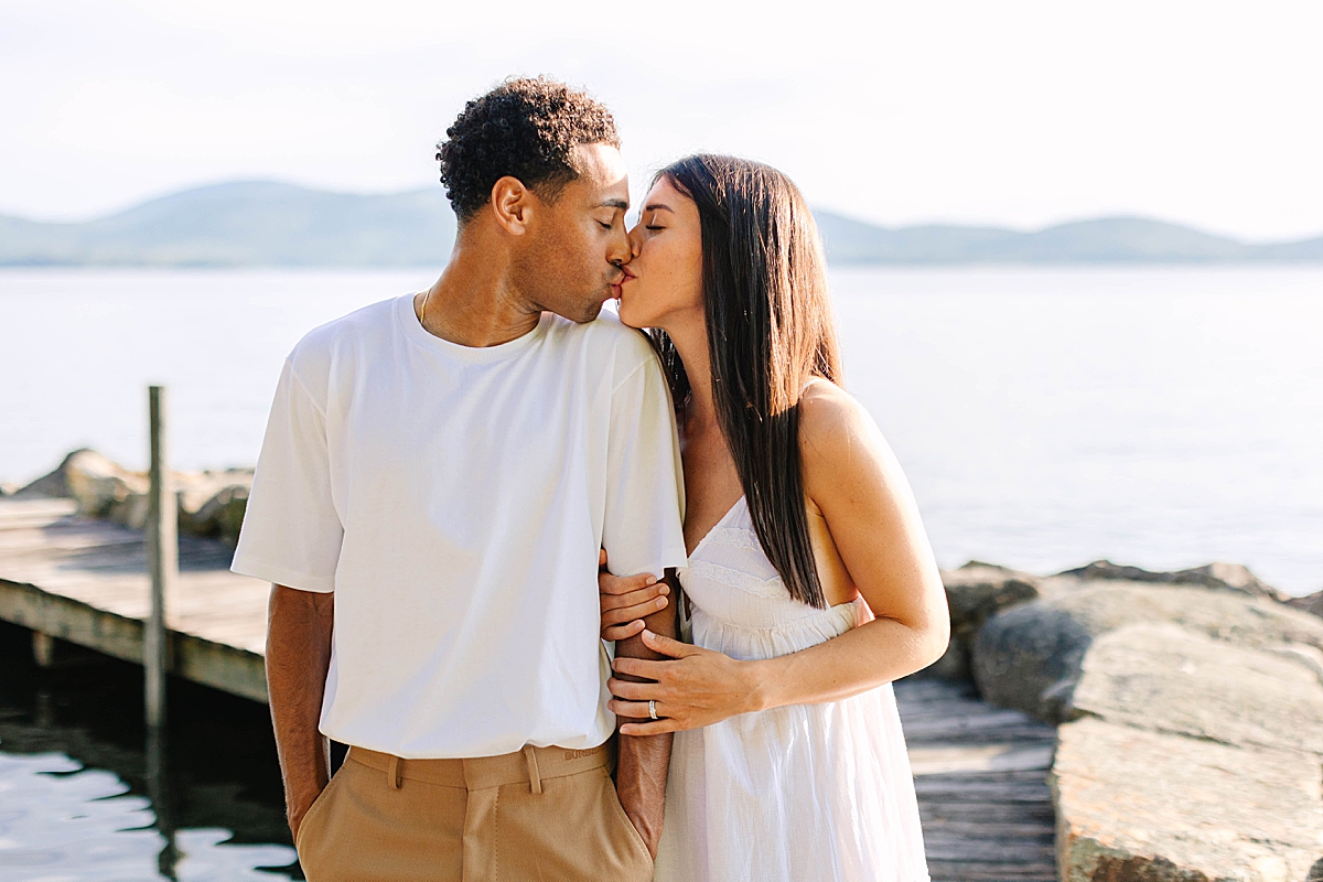 Couple sharing a kiss while standing on a wooden dock, with lake water and hills in the background.