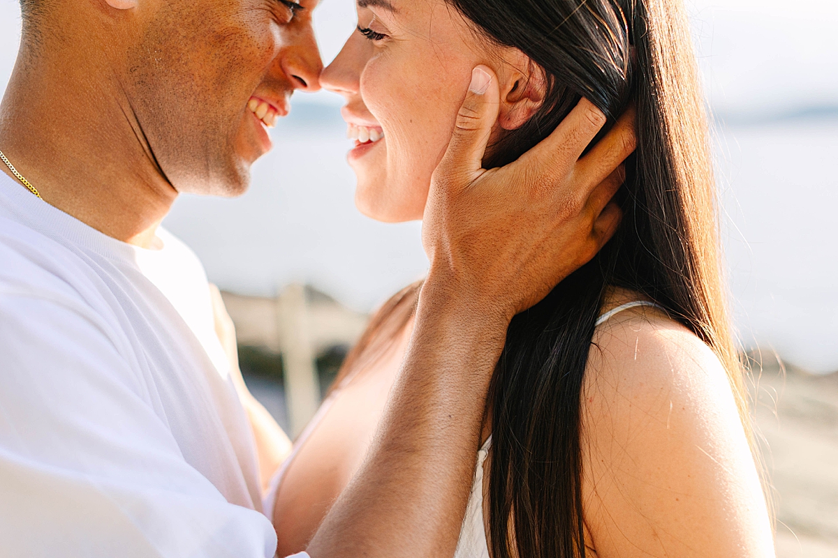 Close-up of a couple smiling face to face, with the man gently cupping the woman’s face near the lake.