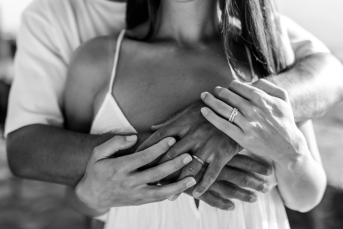 Black and white close-up of a couple’s intertwined hands resting over the woman’s chest, showing wedding rings.
