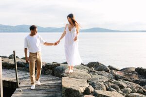 Man helping a barefoot pregnant woman balance on rocks near a dock, with calm water and hills in the background.