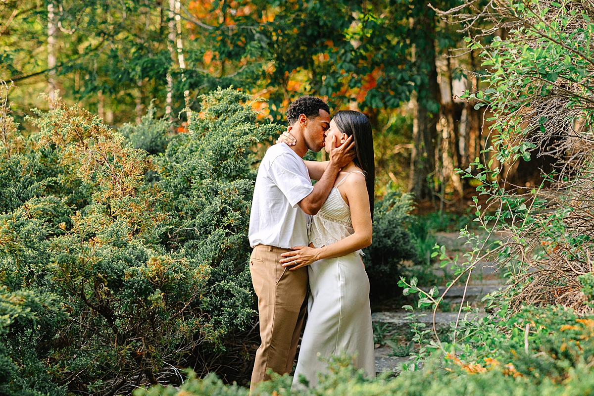 Couple kissing among tall shrubs and trees, with the woman resting her hand on the man's chest.