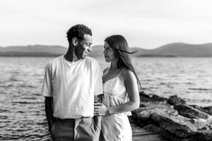 Black and white image of a couple gazing at each other while standing on a dock with the lake behind them.