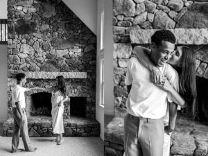 Black and white image of a couple dancing barefoot inside a cozy home, in front of a large stone fireplace.