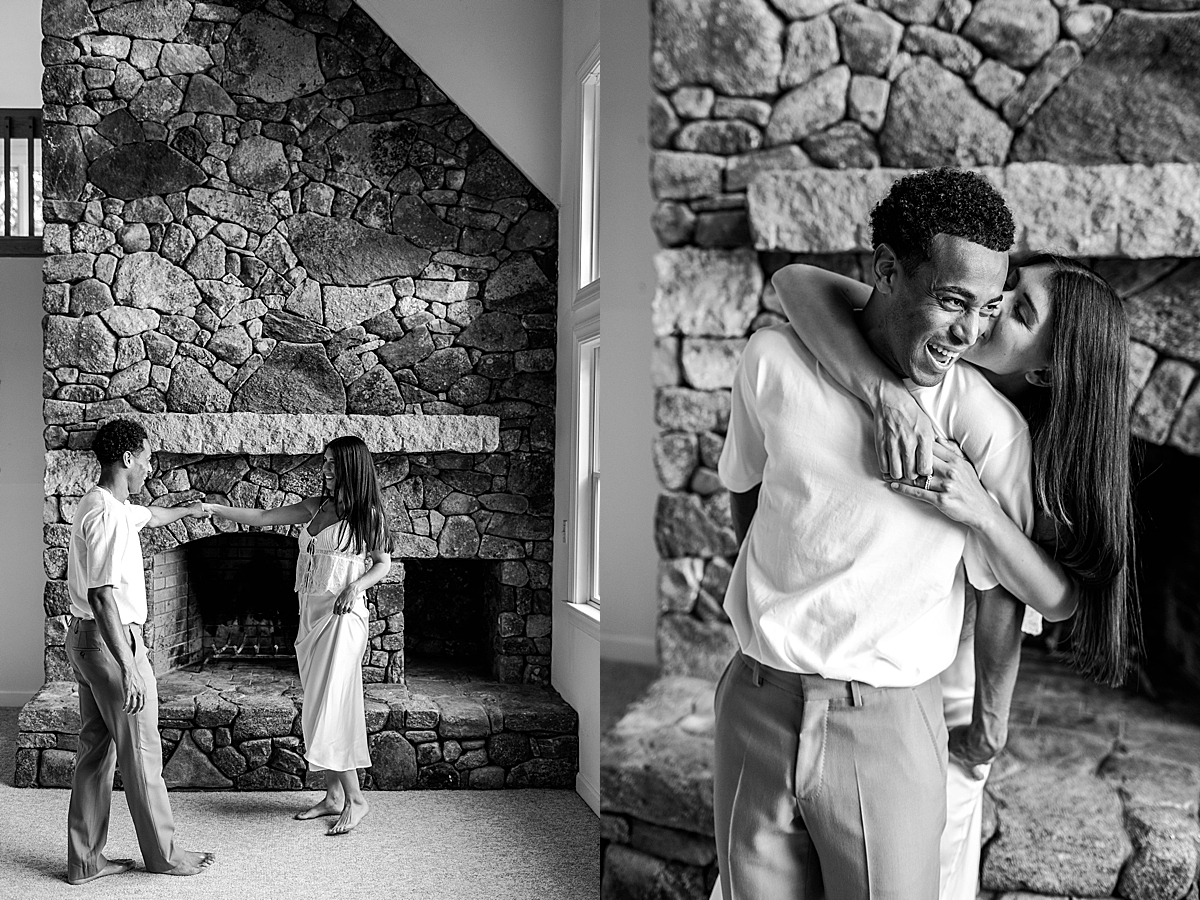 Black and white image of a couple dancing barefoot inside a cozy home, in front of a large stone fireplace.