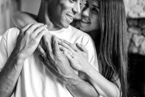 Black and white close-up of a couple embracing indoors, with their hands intertwined and wedding rings visible.